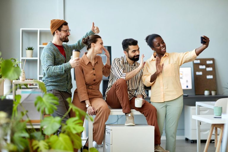 Side view of four young cheerful employees waving hands and looking at smartphone camera with smiles