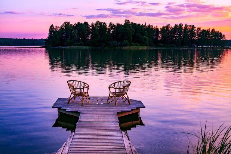 Two rattan wooden chairs and glasses of red wine on a pier overlooking a lake at sunset